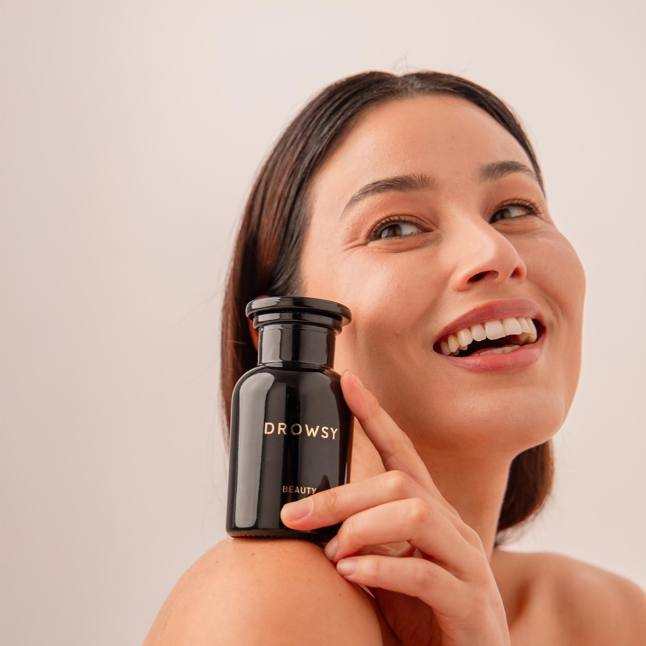 A smiling woman holds a black “Supplements - Beauty” bottle from drowsysleepco near her face, showcasing its skin-enhancing benefits for a youthful complexion against a neutral background.