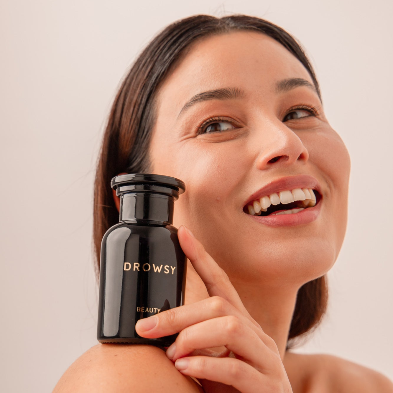 A smiling woman with dark hair holds a black bottle labeled Drowsy Beauty close to her face against a light background.