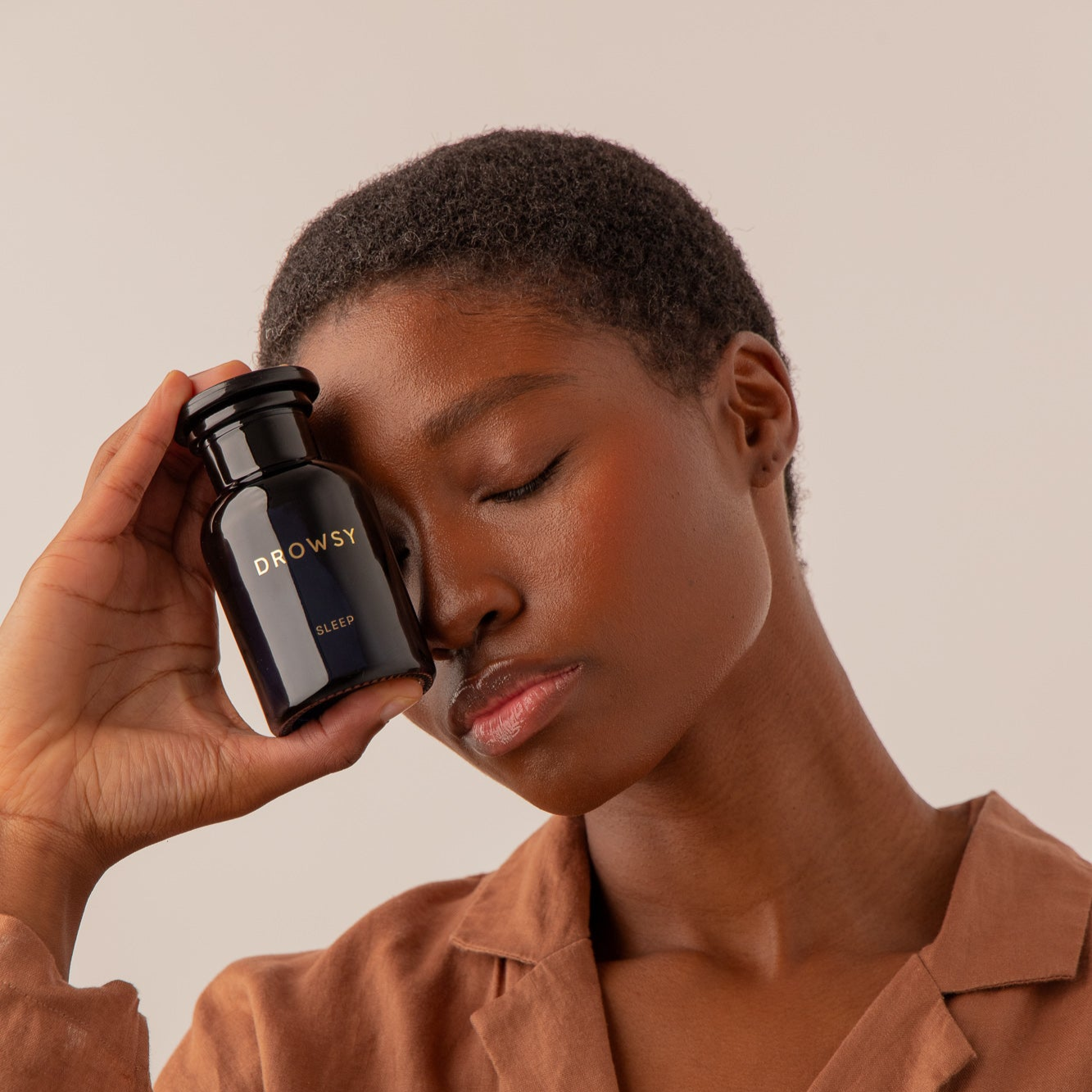 A person with short hair and closed eyes gently holds a black us.drowsysleepco Supplements - Sleep bottle to their face, wearing a brown top against a neutral background; this nightly supplement encourages relaxation.