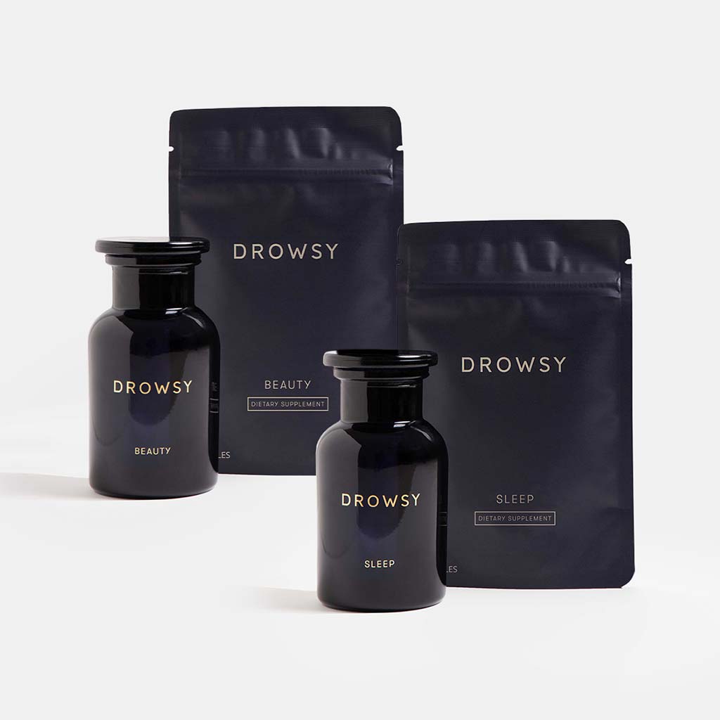 Two dark glass jars and two resealable pouches, all labeled “Drowsy” in gold, from us.drowsysleepco’s Beauty Sleep Clinic supplements. The clean, minimal background highlights their elegant sleep and skin supplement duo presentation.