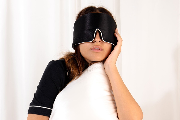 A person wearing a black sleep mask holds a white pillow against their cheek, dressed in dark pajamas with white trim, standing in front of a white background.