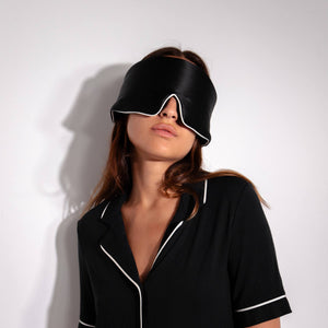 A woman in Black Jade - Pipe Dreams Edition pajamas by us.drowsysleepco stands against a white background, covering her eyes with a large black silk mask for ultimate beauty sleep.