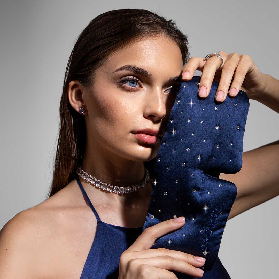 A woman with long brown hair holds the us.drowsysleepco Silk Sleep Eye Mask Crystals by Swarovski®—a navy blue, 100% blackout mask—beside her face. Shes in a dark blue halter dress and crystal jewelry, set against a neutral background.
