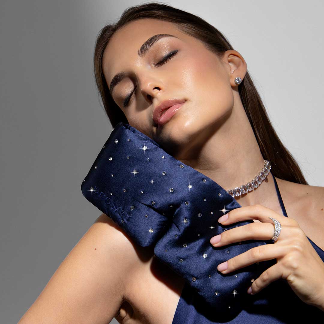 A woman with closed eyes holds the us.drowsysleepco Silk Sleep Eye Mask Crystals by Swarovski®—a navy blue, 100% blackout mask—to her face, wearing a crystal-accented necklace, ring, and earrings and dressed in dark satin.