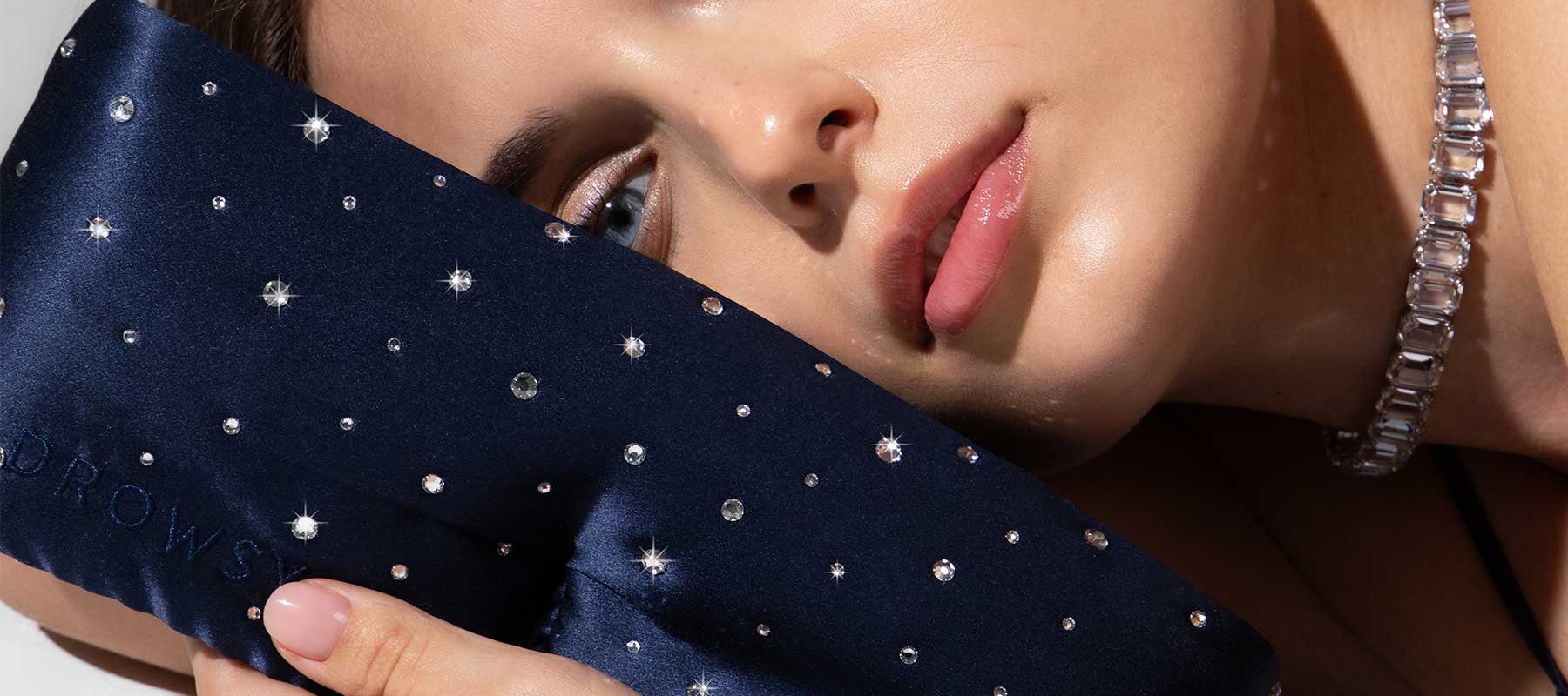 A woman with glossy skin rests her face on a navy blue, jewel-studded eye mask. She wears a sparkly necklace and looks relaxed, with sparkling accents on the mask creating a luxurious feel.