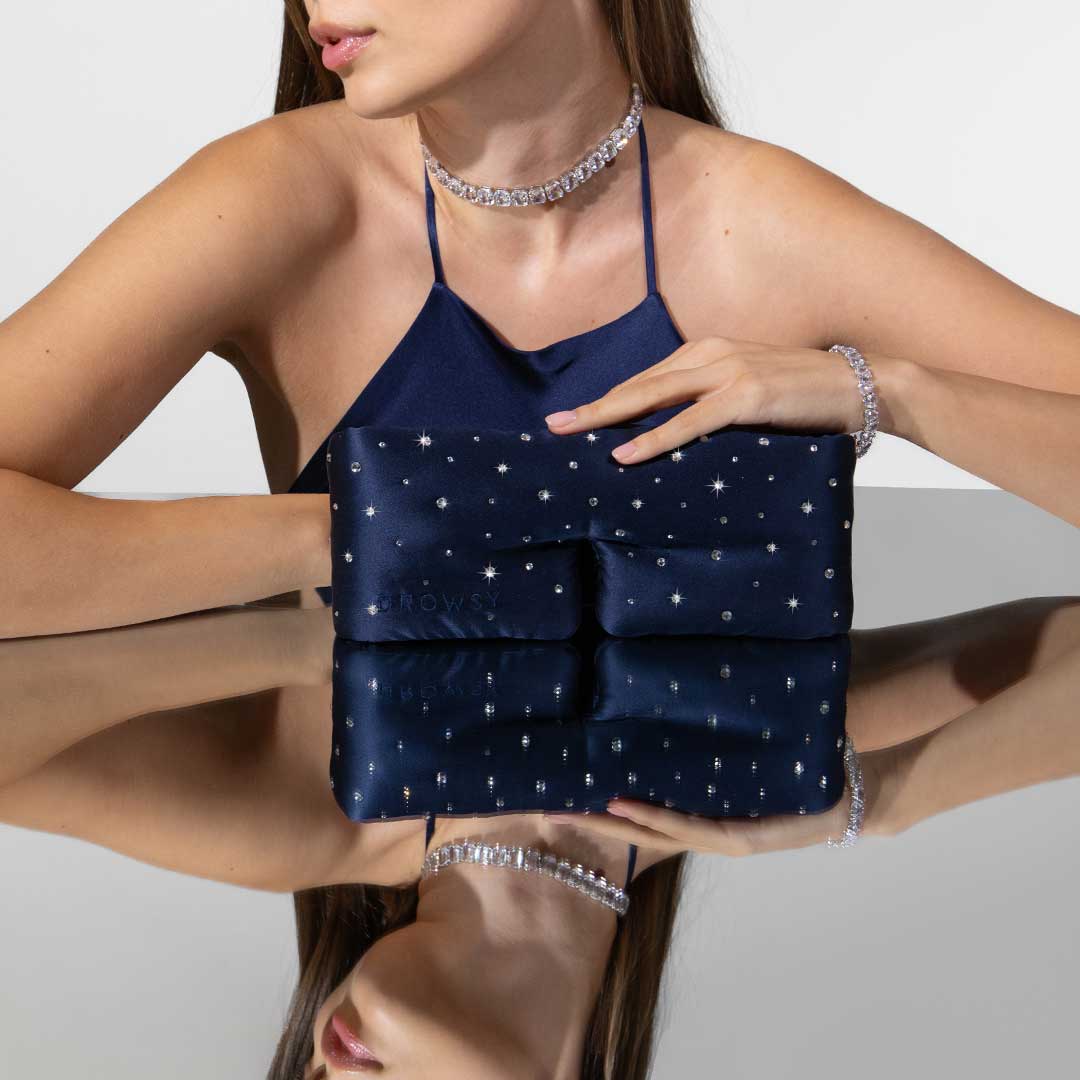 A woman in a navy halter dress and sparkling jewelry sits at a reflective surface, holding the Silk Sleep Eye Mask Crystals by Swarovski® from us.drowsysleepco. Her face is partially visible.