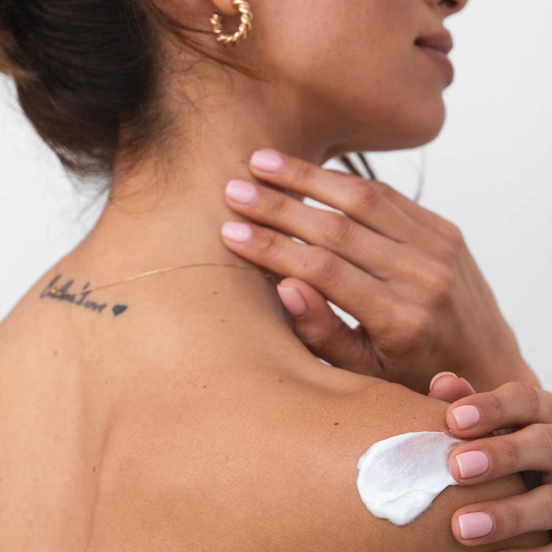 A woman with light pink nails applies us.drowsysleepco Beauty Sleep Body Cream to her shoulder. She has a script and heart tattoo on her upper back and wears a gold hoop earring and delicate gold necklace.