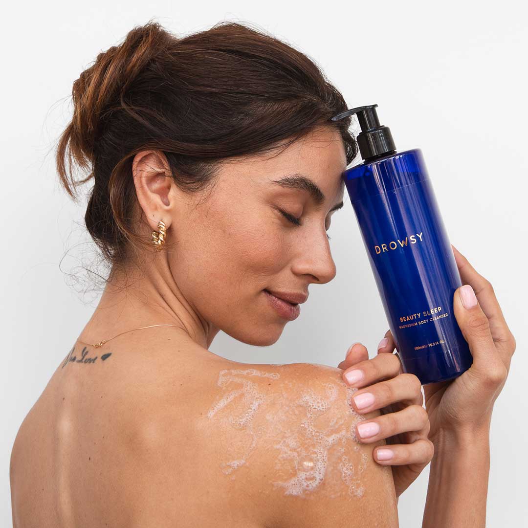 A woman with closed eyes smiles gently, holding a blue bottle of us.drowsysleepco Beauty Sleep Body Cleanser near her face. She has soapy lather on her shoulder and wears gold hoop earrings.