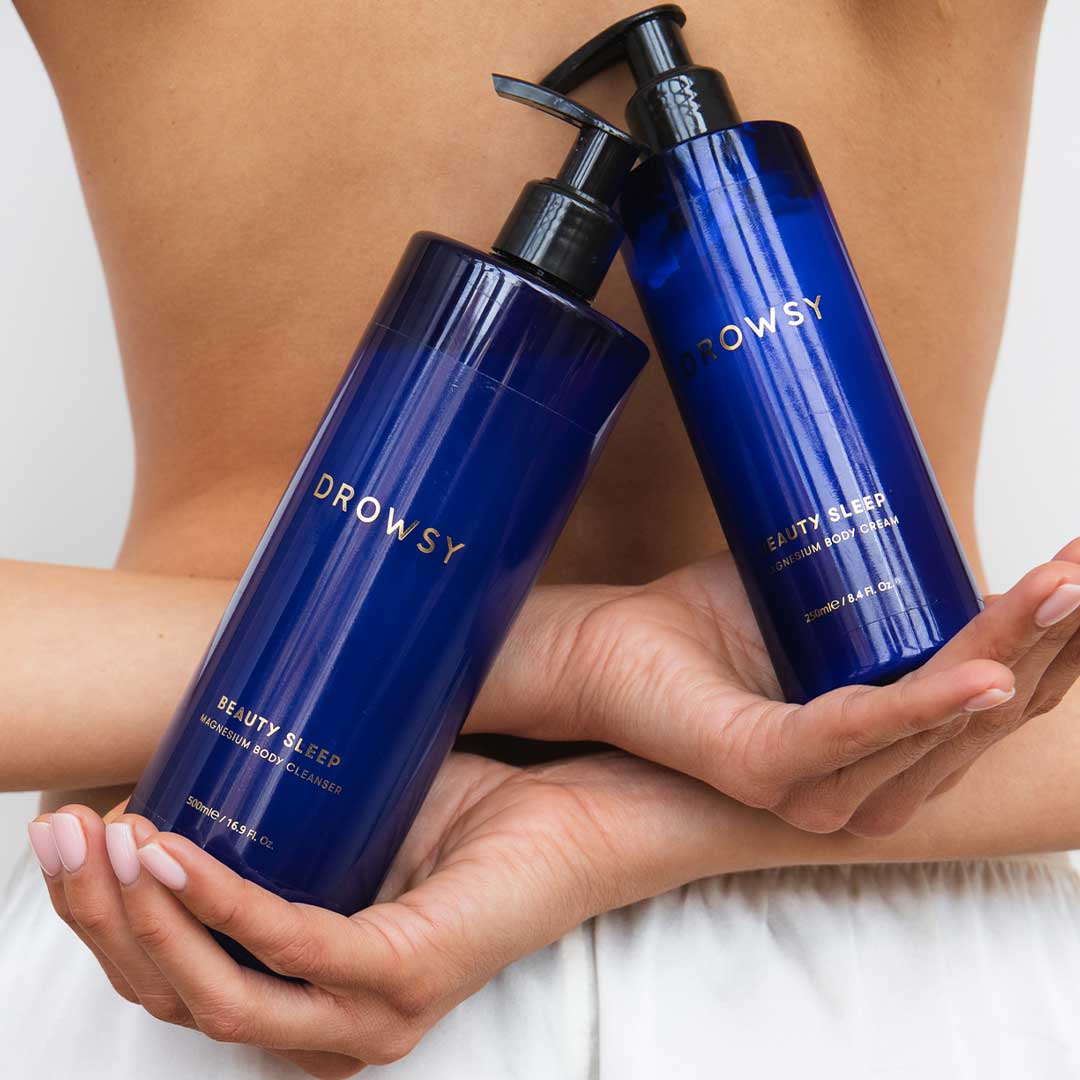 A person in white pants holds two blue bottles of Beauty Sleep Ritual by us.drowsysleepco with pump dispensers, highlighting this magnesium-infused pre-sleep duo against their bare back.