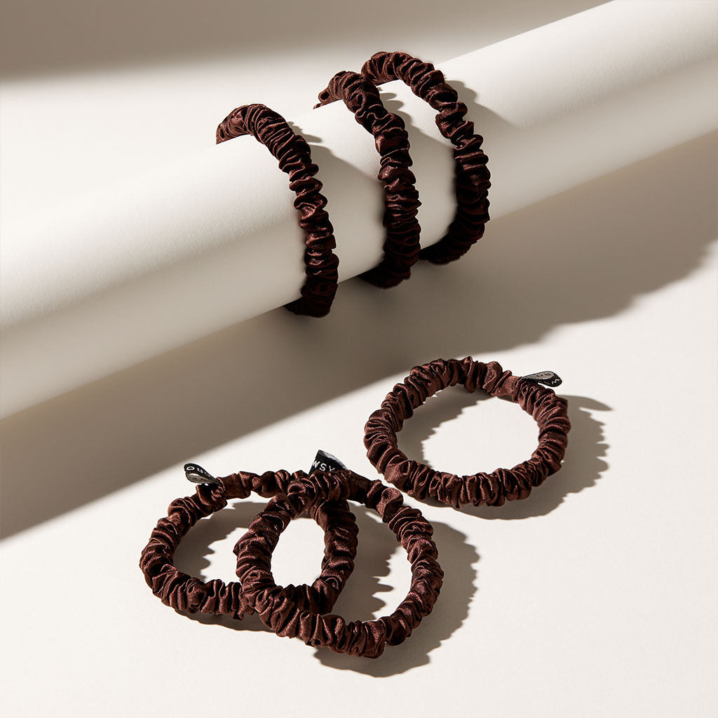 The drowsysleepco Skinny Scrunchie Set - Cocoa Glow features four dark brown silk scrunchies (22 momme), shown on a white cylindrical rod—two dangling, two lying flat below—with soft shadows highlighting their style and hair-friendly design.