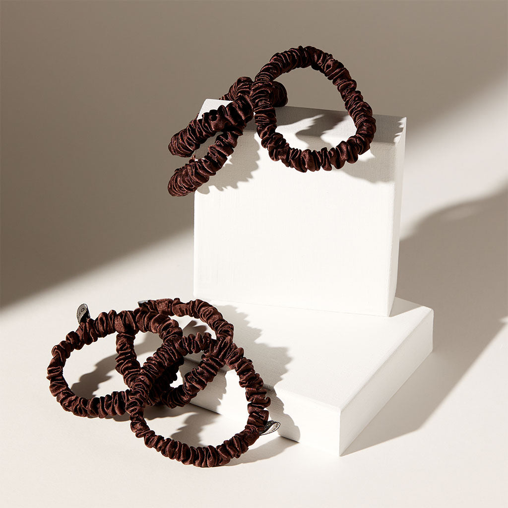The drowsysleepco Skinny Scrunchie Set - Cocoa Glow features four brown, ruffled silk scrunchies (22 momme), elegantly styled on white blocks with soft shadows. Ideal for hair health.