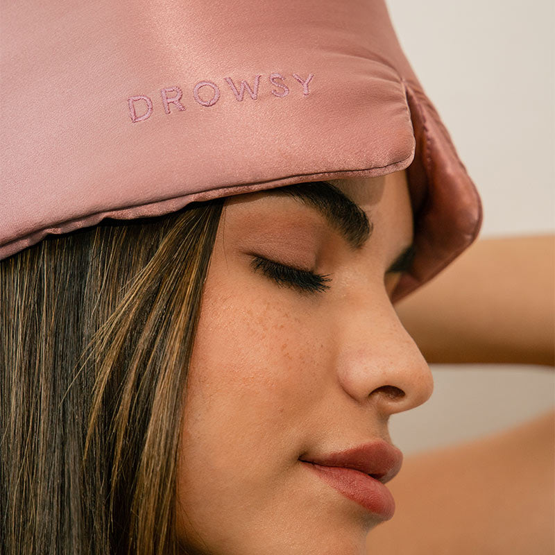 A woman with closed eyes wears a pink satin sleep mask embroidered with the word DROWSY. Her expression is calm and relaxed, and her straight brown hair is visible.