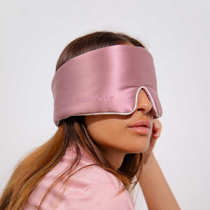 A woman with long brown hair wears the Damask Rose - Pipe Dreams Edition sleep mask by us.drowsysleepco, a shiny pink silk mask with an adjustable strap labeled DROWSY, covering her eyes. She dons a pink top against a white background.
