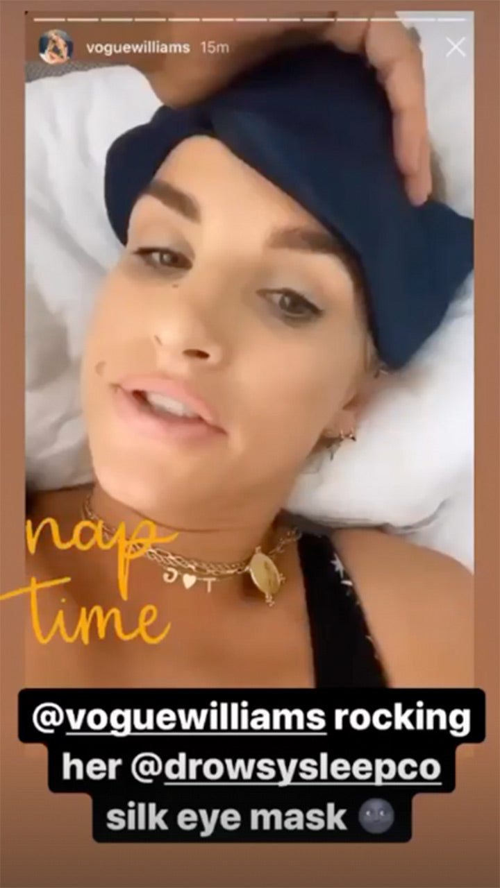 A woman lies on a bed wearing a dark blue silk eye mask pushed up on her forehead. Text reads nap time and mentions @voguewilliams rocking her @drowsysleepco silk eye mask.