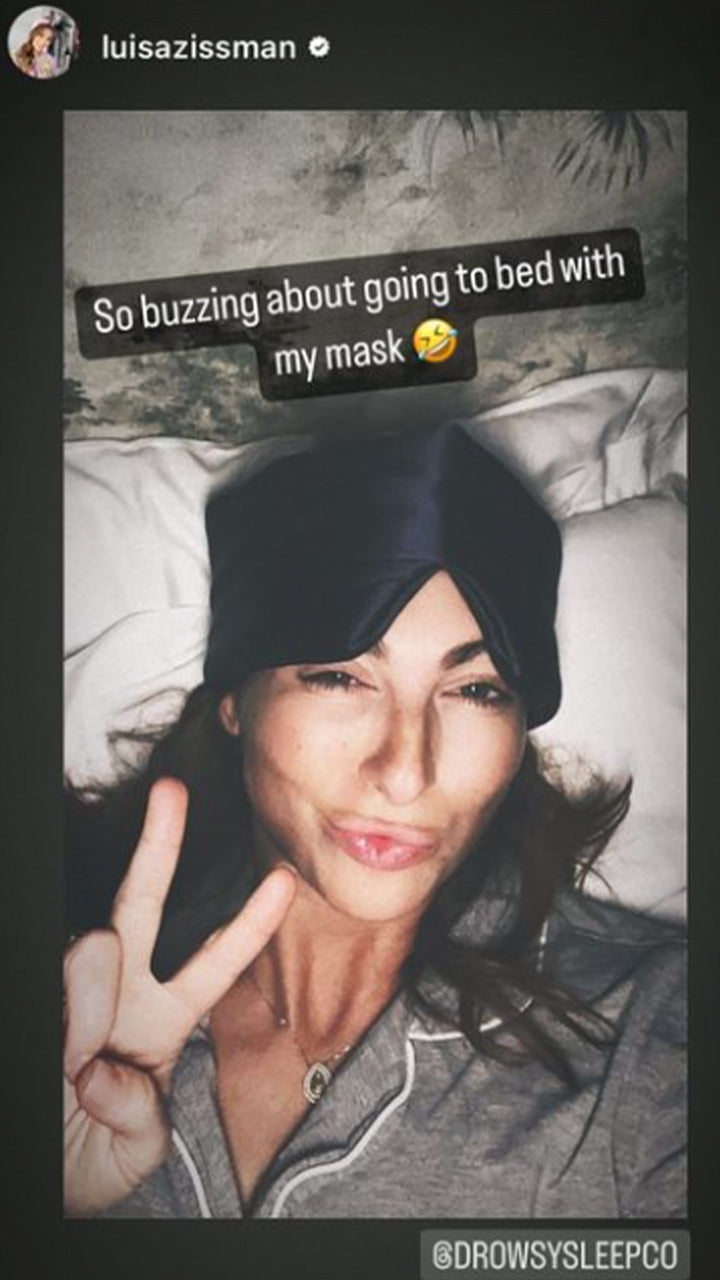 A woman lying in bed wearing a black sleep mask on her forehead, making a peace sign with her hand and a playful face. Text on the image reads, So buzzing about going to bed with my mask 🤣.