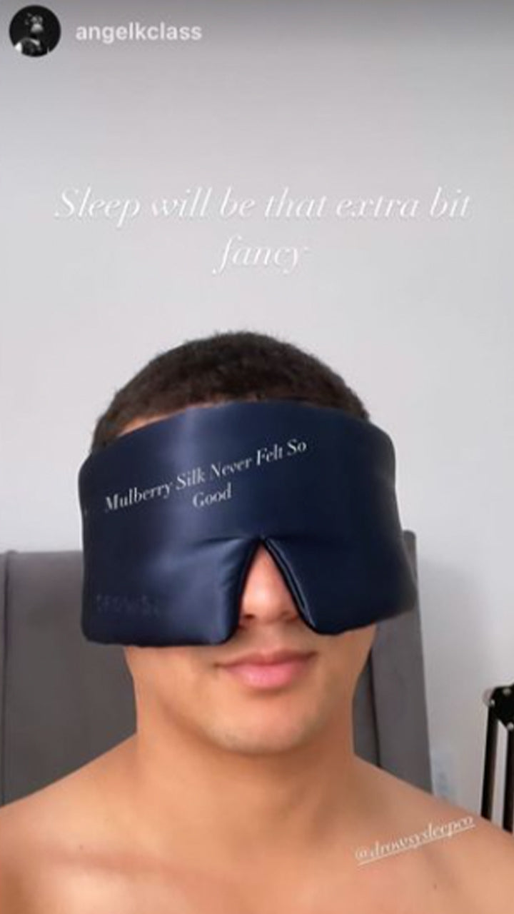 A person wearing a large navy blue silk sleep mask with the words Mulberry Silk Never Felt So Good printed on it. Text above reads, Sleep will be that extra bit fancy. The person is indoors and shirtless.