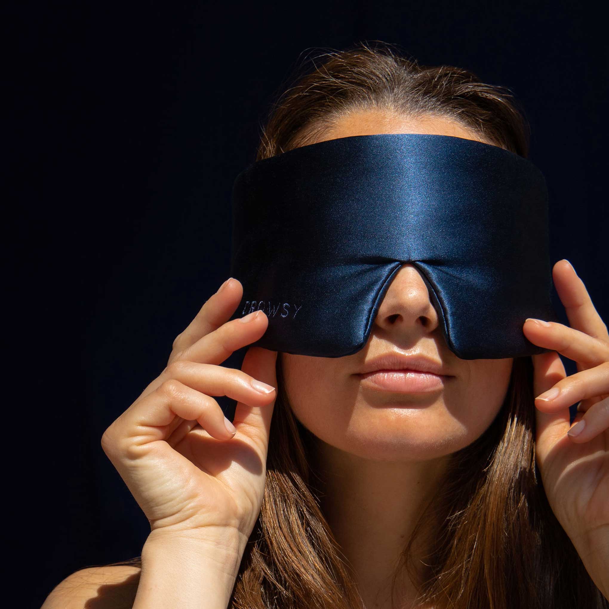 A woman with long brown hair holds a large, dark blue silk sleep mask over her eyes, against a dark background. The mask covers her eyes completely, and she is gently adjusting it with both hands.
