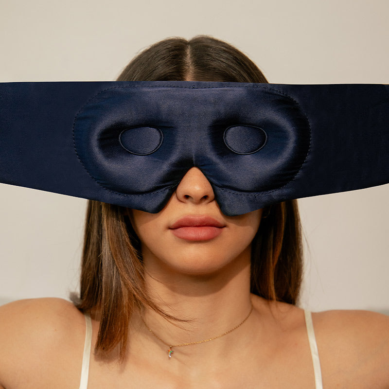 A woman with straight brown hair wears a dark blue silk eye mask with large oval cutouts over her eyes. She has a neutral expression and is dressed in a white camisole. The background is plain and light-colored.