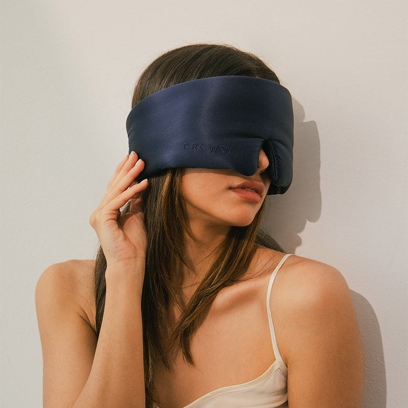 A woman in a white camisole sits against a beige wall, wearing a large, dark blue sleep mask that covers her eyes and nose. She touches the side of the mask with one hand.