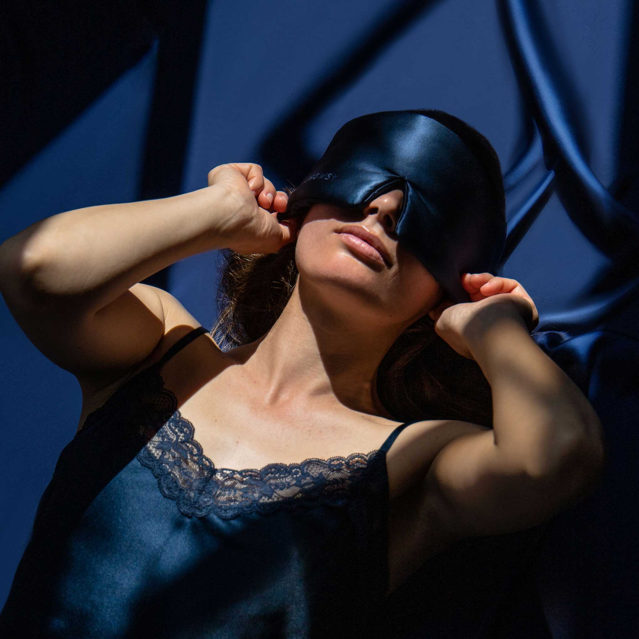 A woman wearing a dark satin sleep mask and navy lace-trimmed camisole stands in dramatic lighting, adjusting the mask over her eyes. Blue satin fabric drapes in the background.