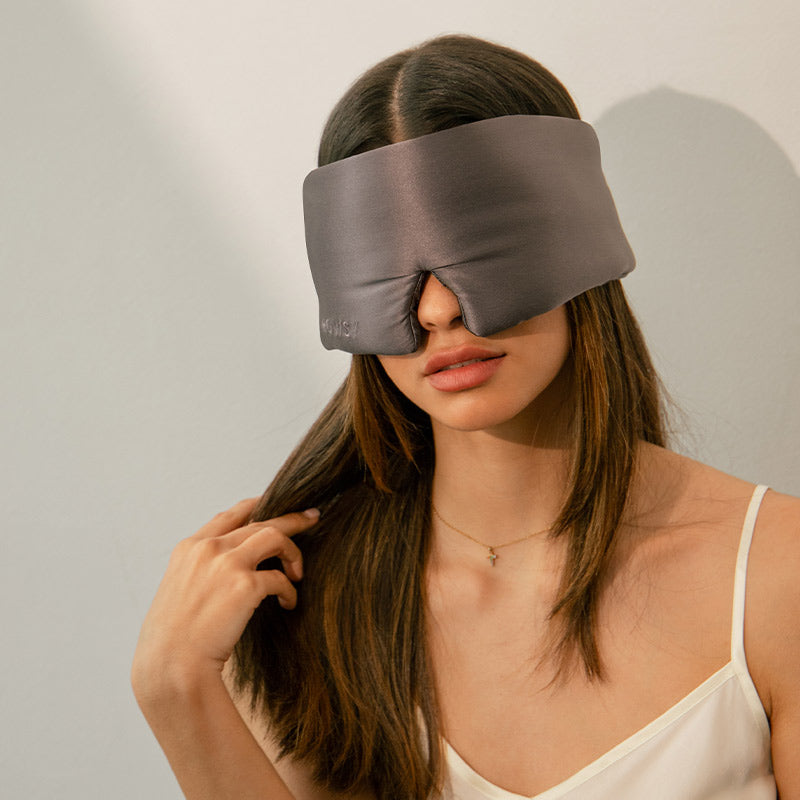 A woman with straight brown hair wears a padded dark grey sleep mask that covers her eyes and nose. She is dressed in a white camisole and is touching her hair with one hand, sitting against a light-colored wall.
