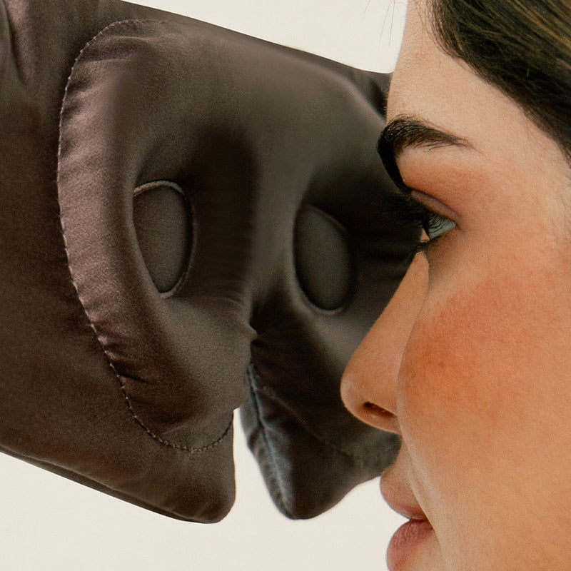 A close-up of a womans face wearing a brown satin sleep mask with large eye cavities, allowing space around her eyes and eyelashes.