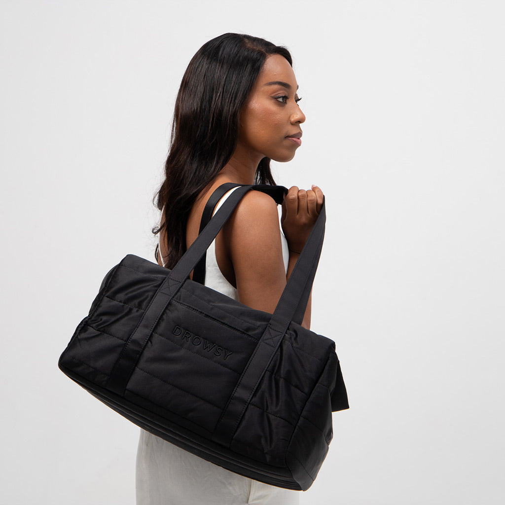 A woman with long dark hair in a white sleeveless top stands in profile, holding the Drowsy Silk Weekender - Black Jade duffel bag with DROWSY embroidery, perfect for essentials, against a plain white background.