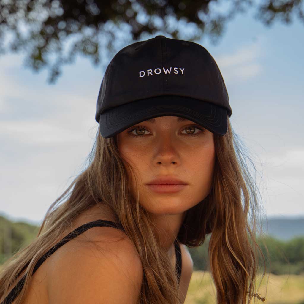 A woman with long brown hair wears the Drowsy Silk Cap - Black Jade by Drowsy and a black strap top, gazing at the camera outdoors on a sunny day with blurred trees and sky in the background.
