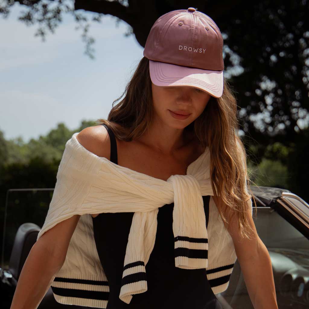 A woman in the Drowsy Silk Cap - Damask Rose by Drowsy, a black tank top, and a white sweater over her shoulders stands beside a convertible car on a perfect sunny day.