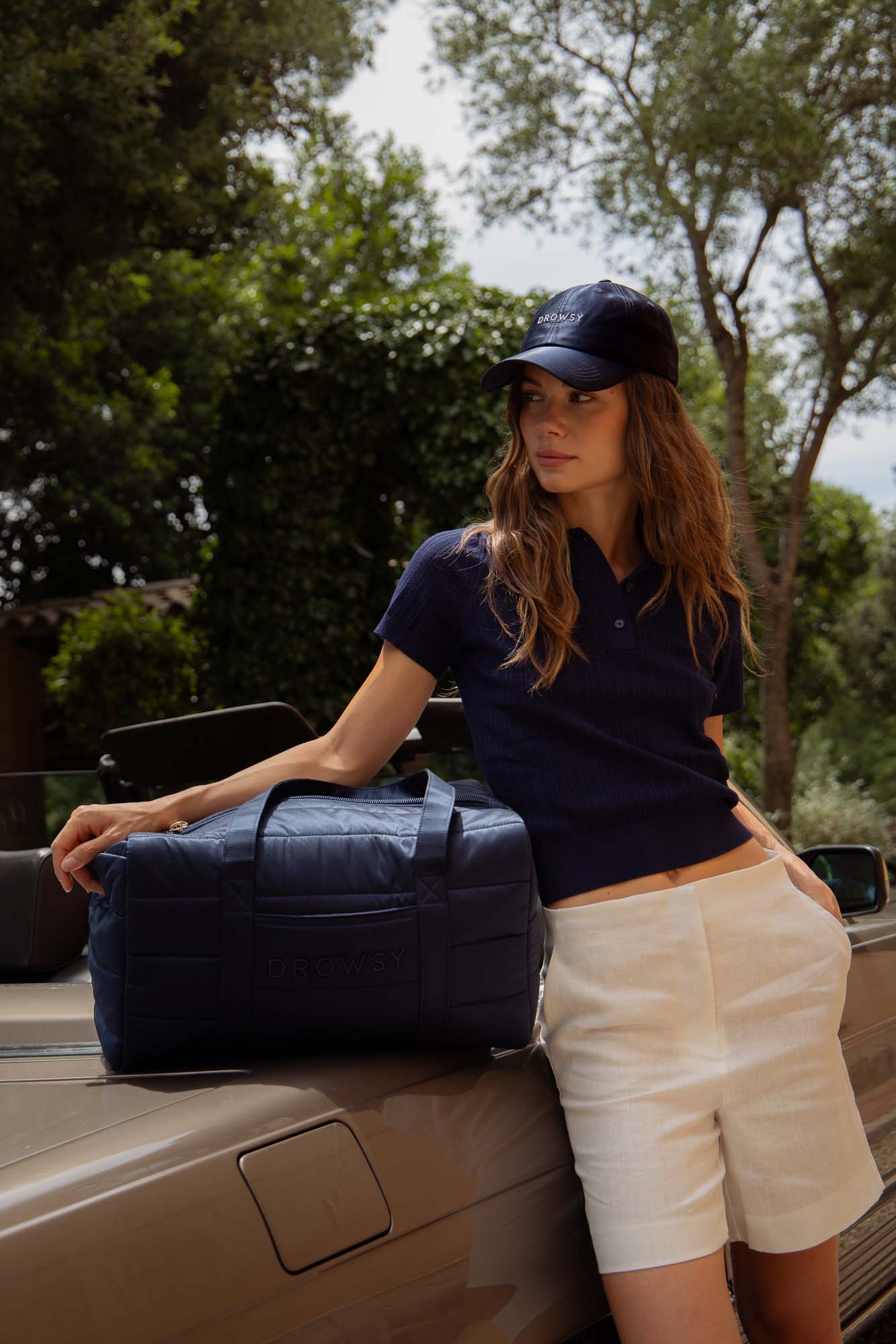 A woman in a navy polo shirt, white shorts, and a navy cap leans against a car with a large navy duffel bag. Trees and greenery are visible in the background on a sunny day.