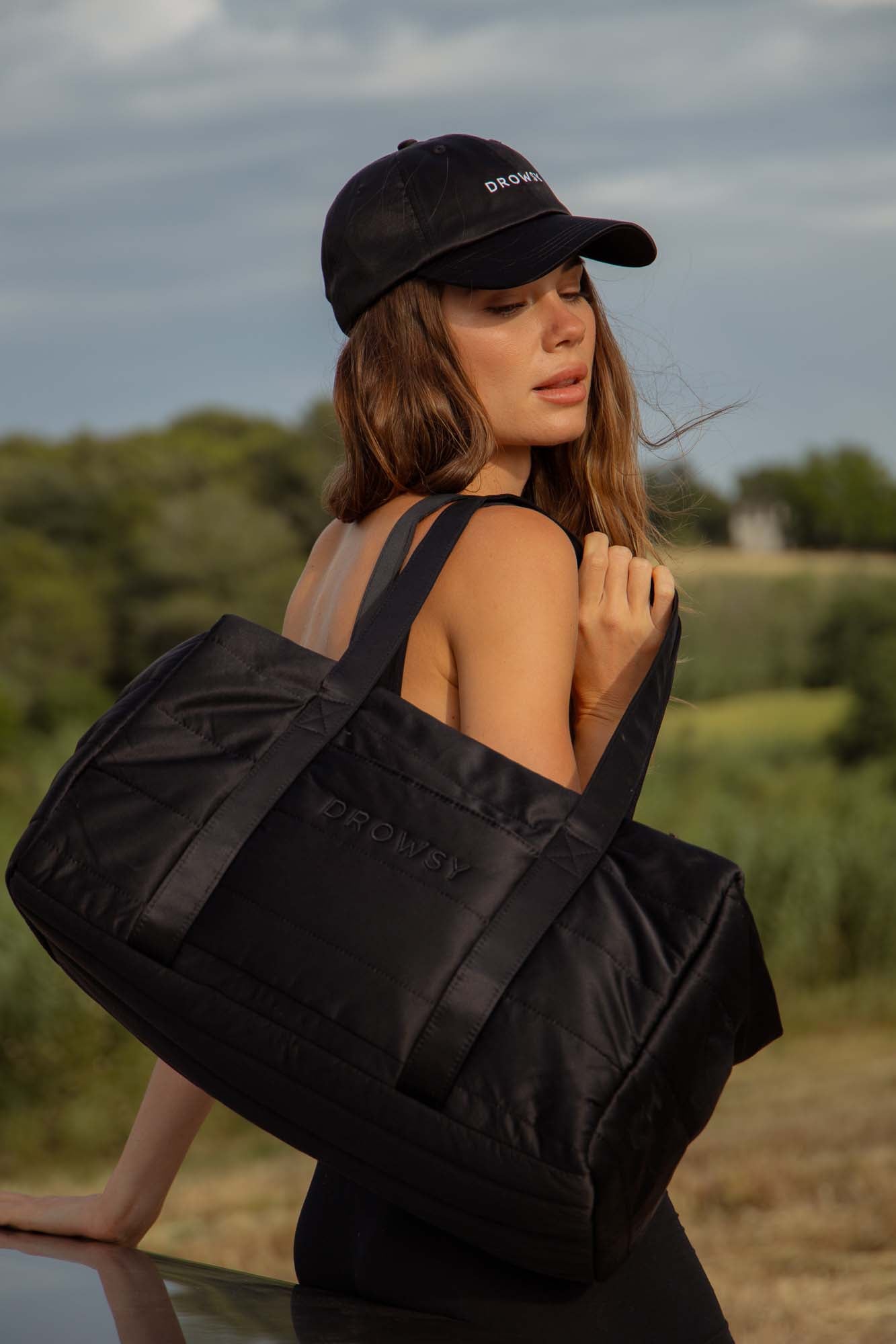 A woman wearing a black cap and sleeveless top carries a large black duffel bag over her shoulder. She is outdoors in a grassy, rural area with trees and a cloudy sky in the background.