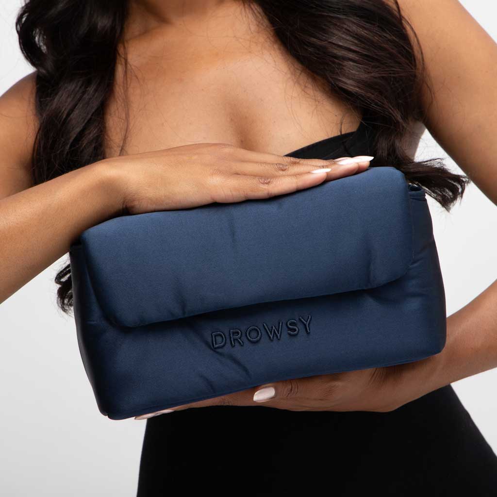 A woman with long dark hair holds the Drowsy Weekend Collection - Midnight Blue + Damask Rose pouch from Drowsy, featuring “DROWSY” embroidered in black. She wears a strapless black top, with one arm around the bag and the other resting on top.