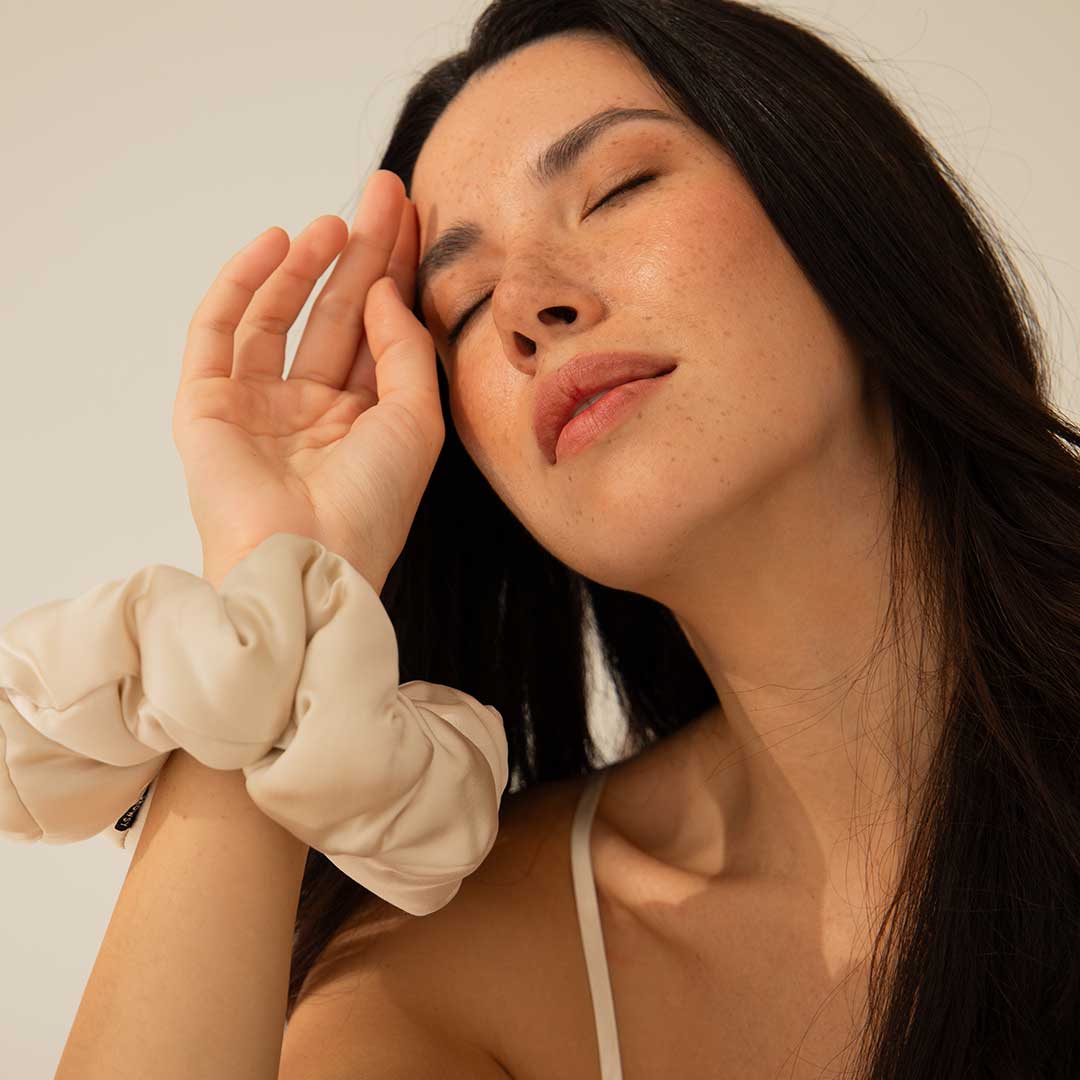 Woman with long dark hair and light skin, eyes closed, touching her forehead with her hand. She wears a cream-colored scrunchie on her wrist and a thin strap top, appearing relaxed and serene in soft lighting.
