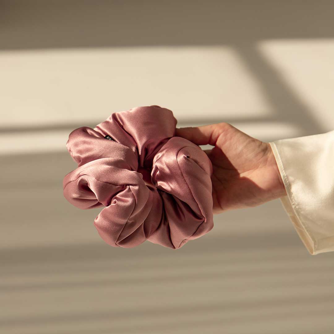 A hand in a white sleeve holds the us.drowsysleepco Damask Rose - Padded Scrunchie, crafted from shiny pink satin to enhance hair health, shown against a softly lit background with gentle shadows.