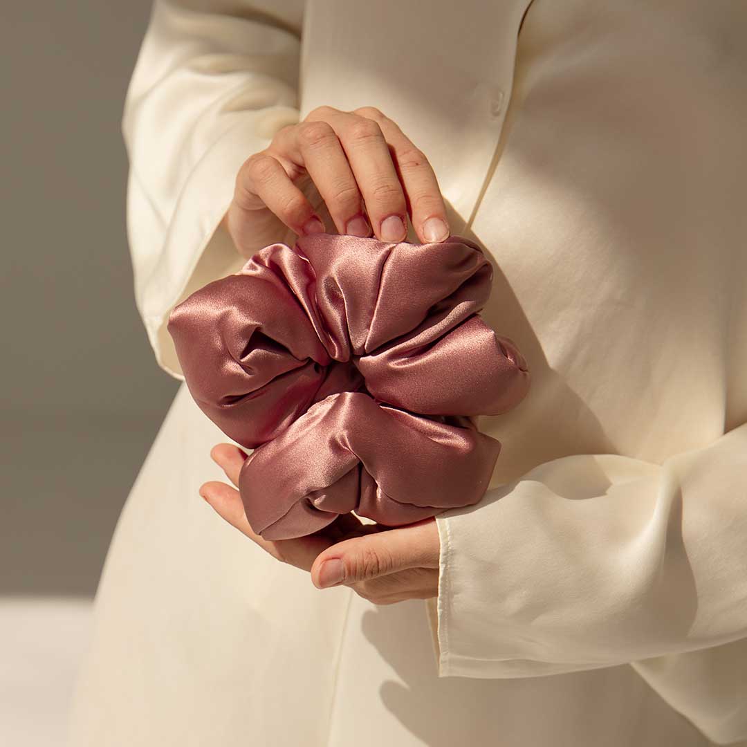 A person in a cream-colored outfit holds a large, shiny pink satin scrunchie with both hands, against a neutral background.