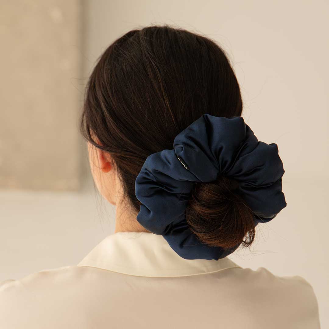 A woman with dark hair in a low bun wears the us.drowsysleepco Midnight Blue - Padded Scrunchie, promoting hair health. She is seen from behind in a cream blouse against a softly blurred background.