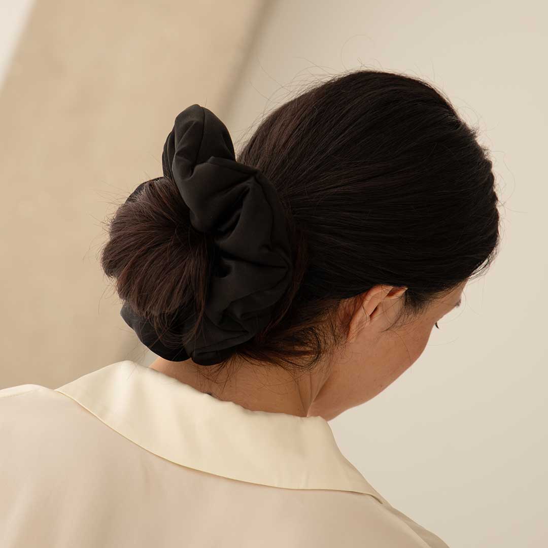 A woman with dark hair in a low bun, secured with the us.drowsysleepco Black Jade - Padded Scrunchie, wears a cream collared shirt and faces a neutral background. The silk-padded scrunchie helps promote hair health.