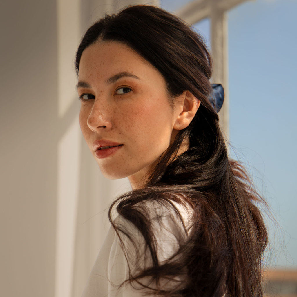 A woman with long dark hair and freckles, wearing a white top, looks over her shoulder while standing near a sunlit window.