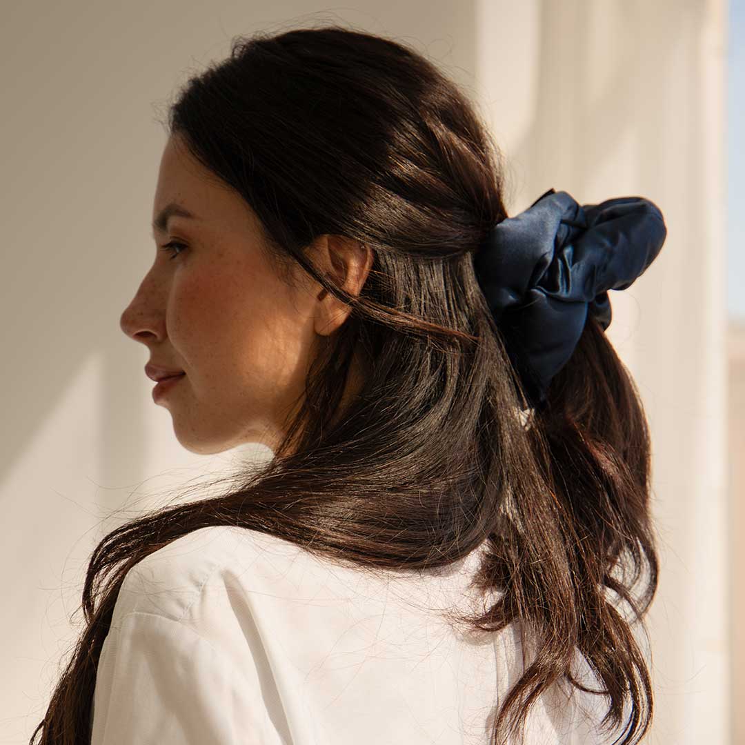 A woman with long brown hair in a half-up style, secured by the Midnight Blue - Padded Scrunchie from us.drowsysleepco, wears a white top and gazes to the side in soft natural light.