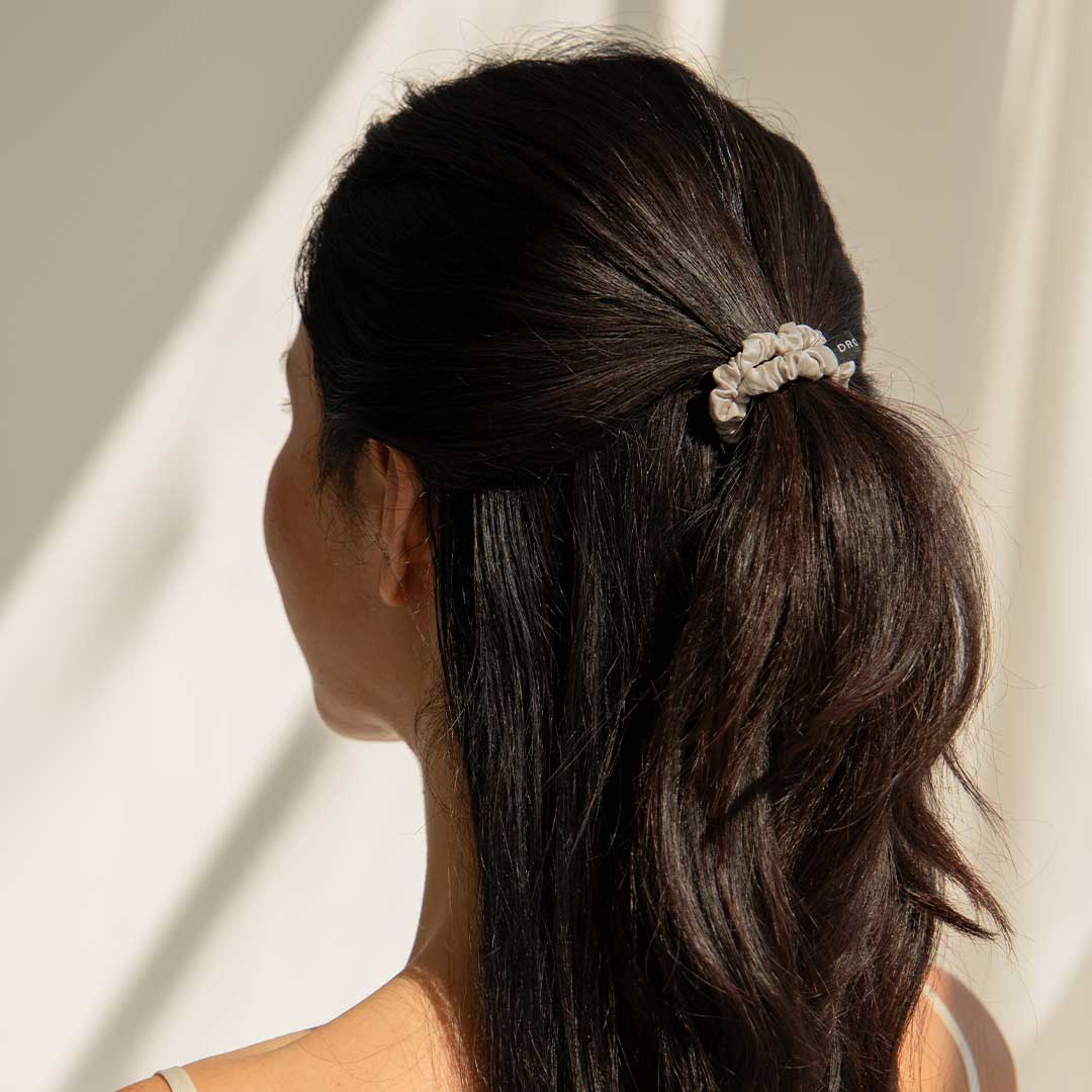 A woman with long dark hair styled in a low ponytail, secured with a light-colored scrunchie, is seen from behind against a softly lit, neutral background.