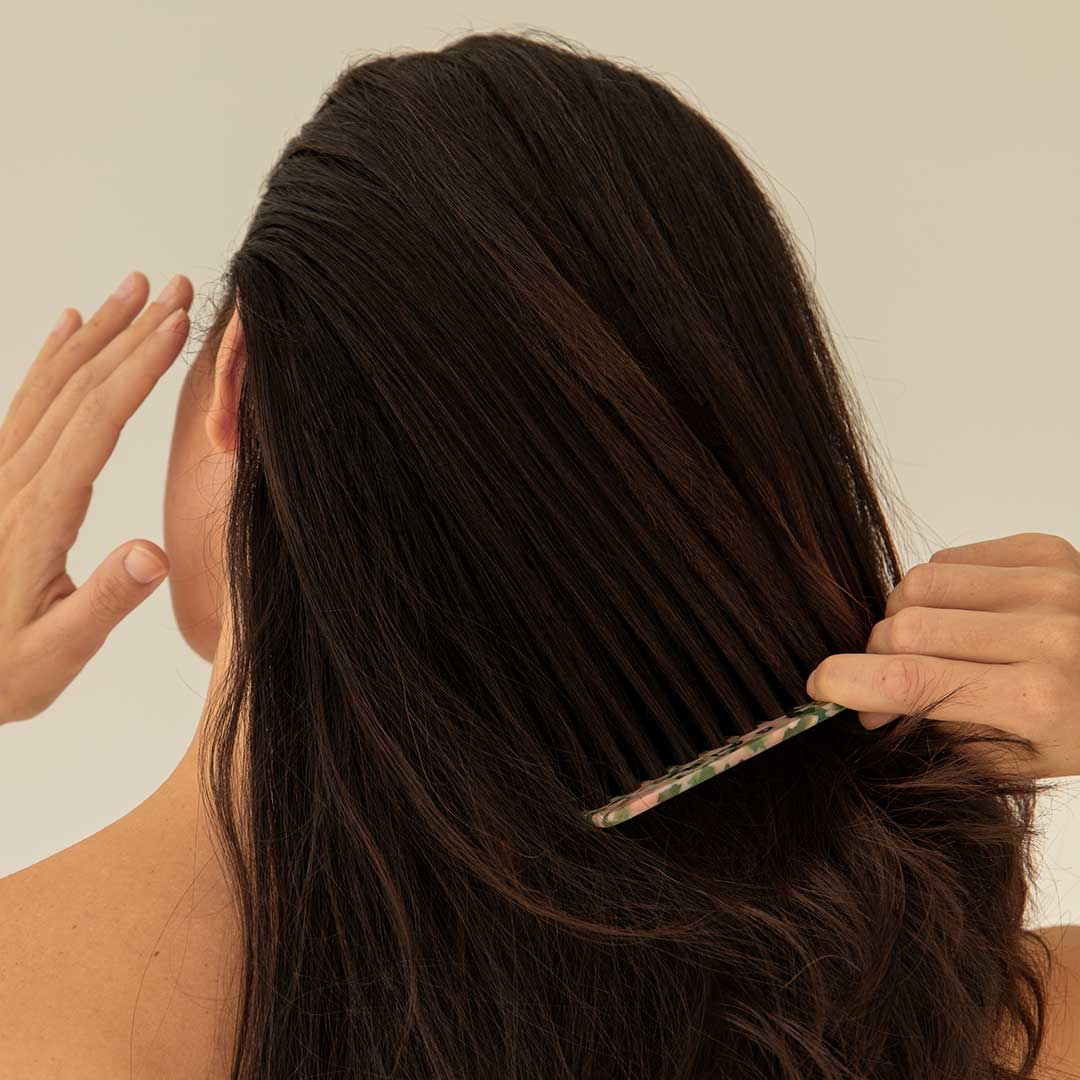 A person with long, dark hair is seen from behind, using a comb to detangle their hair. One hand holds the comb, while the other hand is raised near their head. The background is plain and neutral.