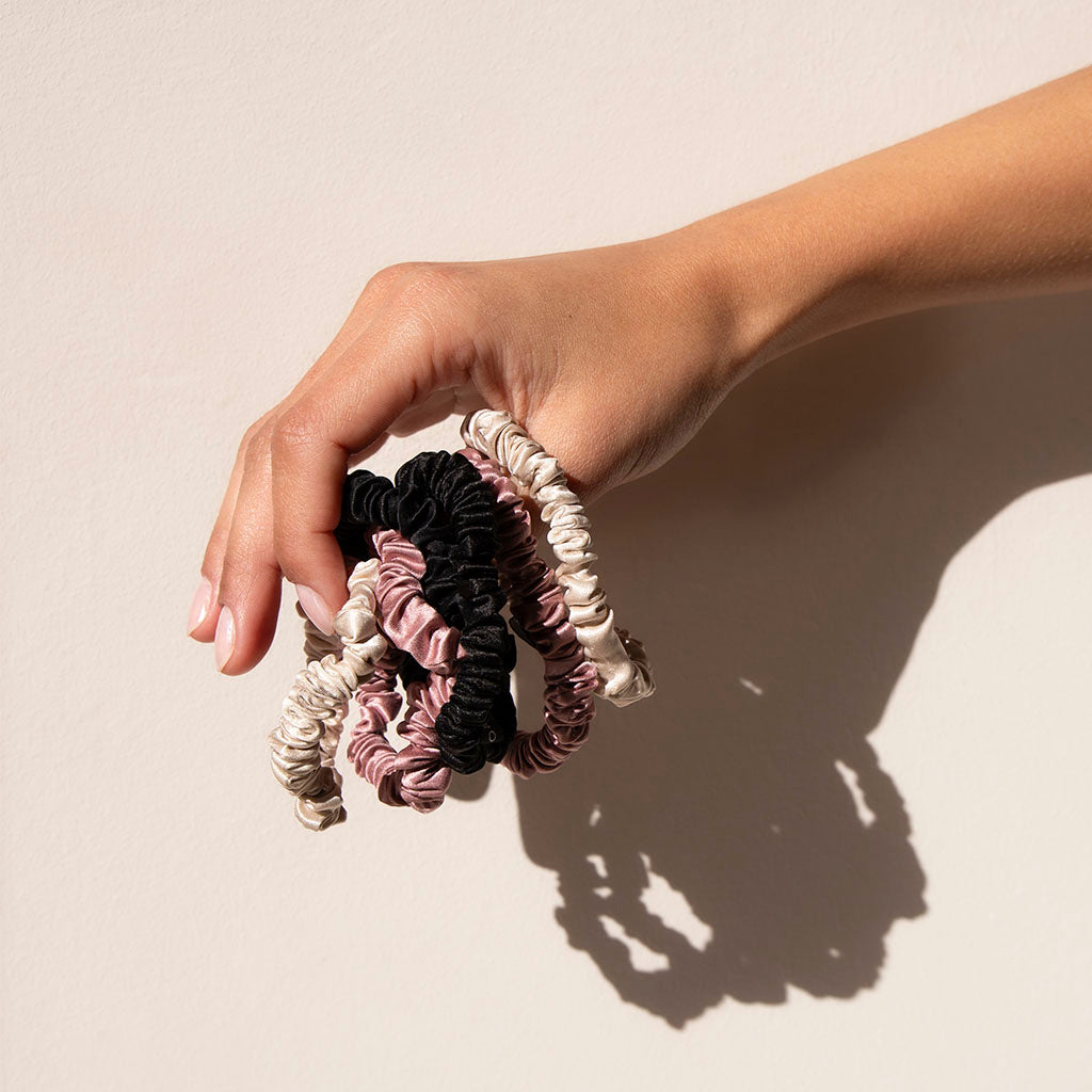 A hand holds the us.drowsysleepco Skinny Scrunchie Set in Rose, Dusty, and Black against a light beige background—ideal for gentle styling and promoting hair health.
