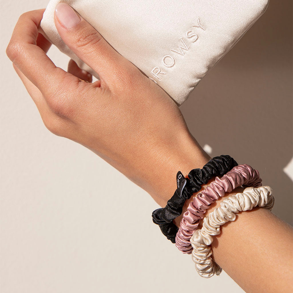 A hand with us.drowsysleepcos Skinny Scrunchie Set—Rose, Dusty, Black (22 momme silk) holds a white satin ROWSY pouch, perfect for stylishly elevating hair health.