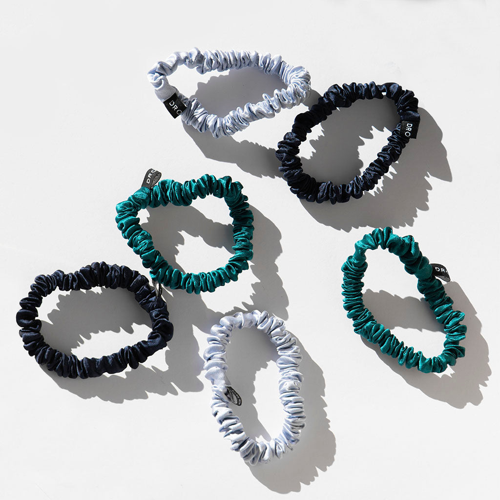 The us.drowsysleepco Skinny Scrunchie Set - Blue & Green Shades includes six 22 momme silk hair ties in black, teal, and light blue, each with a small black tag—ideal for supporting hair health. Displayed on a white surface.