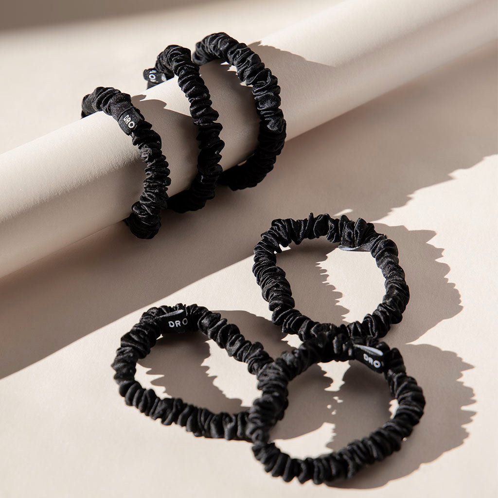 Four Skinny Scrunchie Set - Black Jade by us.drowsysleepco silk scrunchies are displayed on a neutral surface in natural light—two draped over a cylinder, two lying flat—highlighting their hair-friendly benefits.