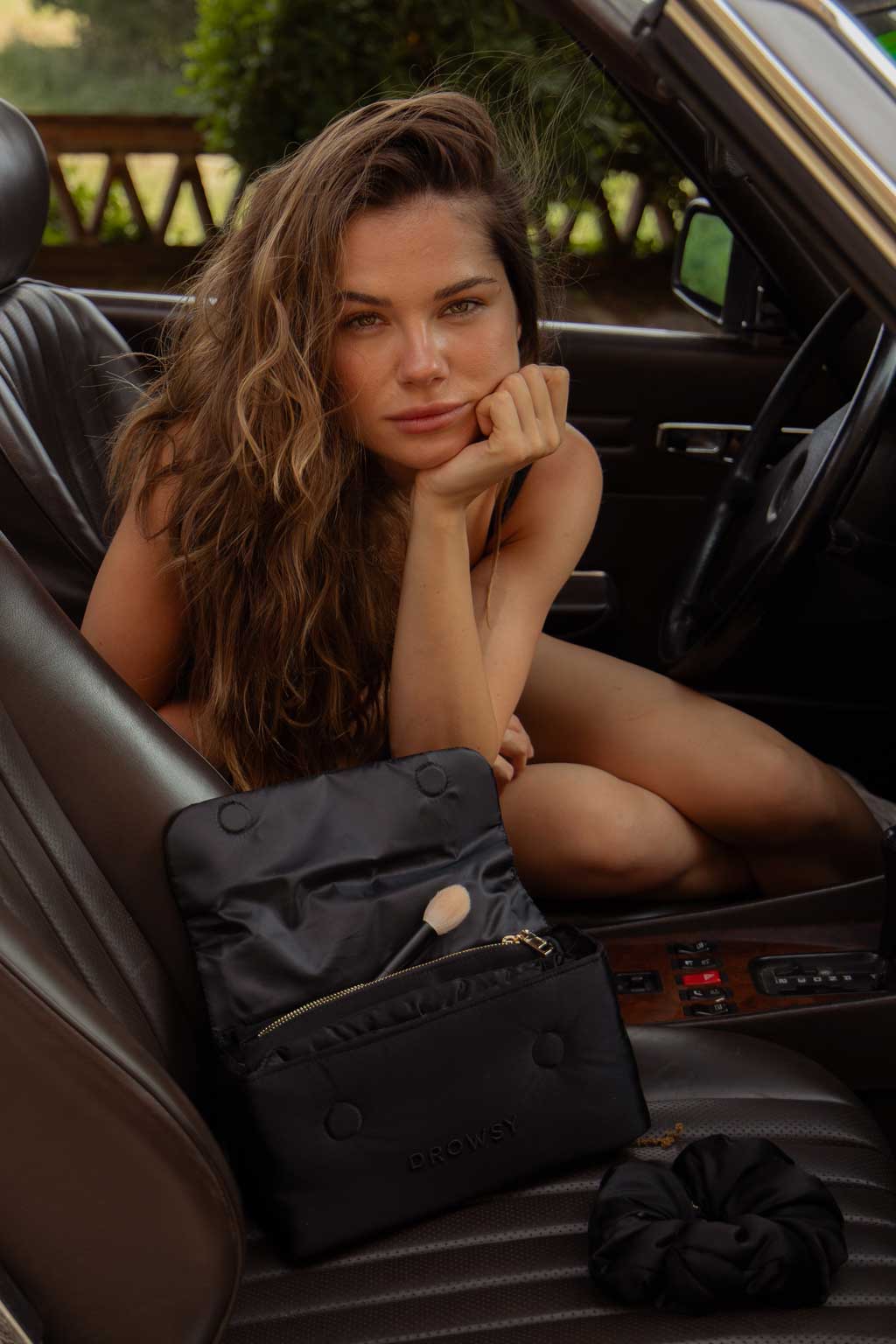A woman with long, wavy hair sits in the driver’s seat of a car, resting her chin on her hand. An open black makeup bag and a scrunchie are on the car seat beside her.