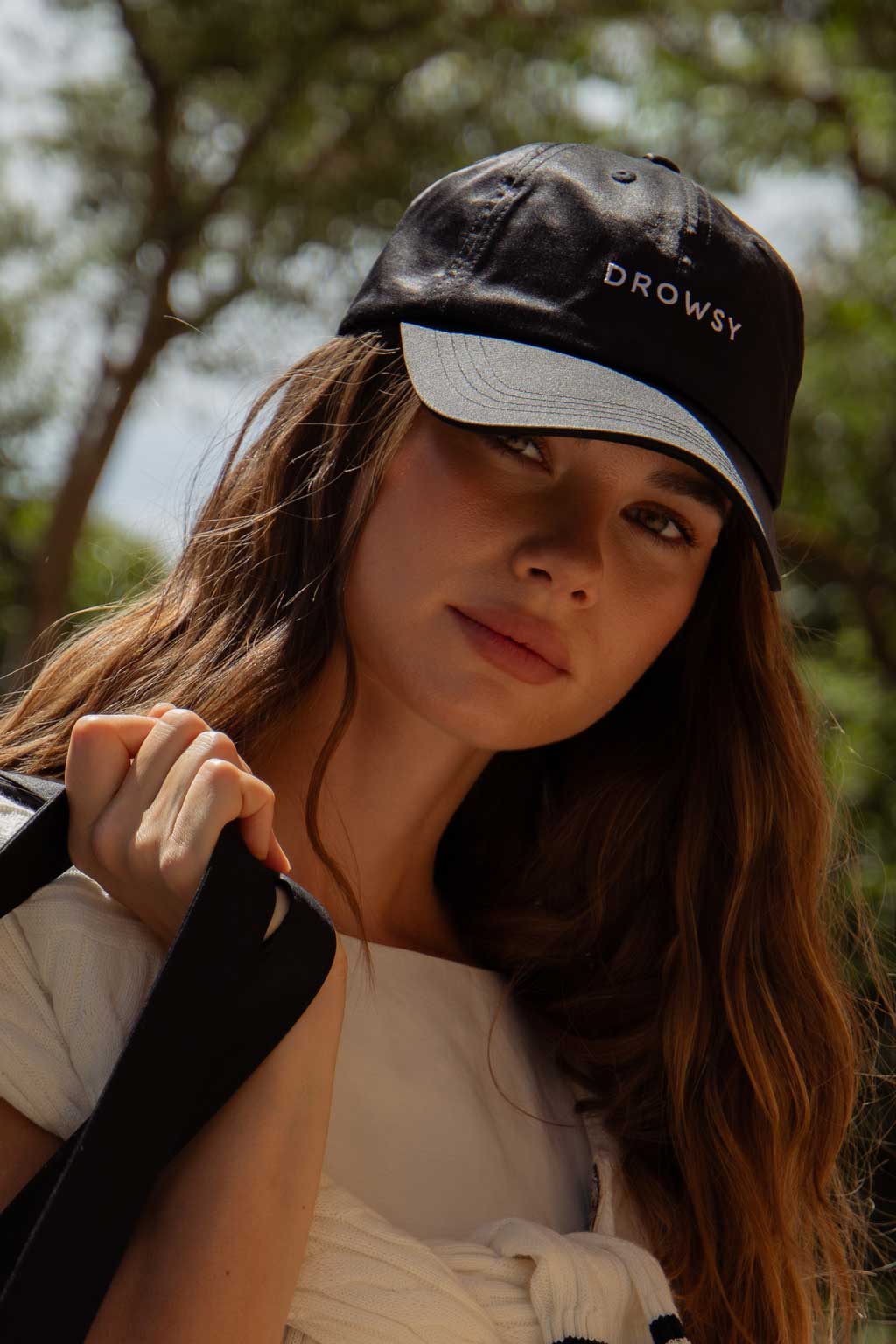A woman with wavy brown hair wearing a black cap with the word DROWSY embroidered on it stands outdoors, holding a black strap over her shoulder and looking calmly at the camera. Trees are blurred in the background.