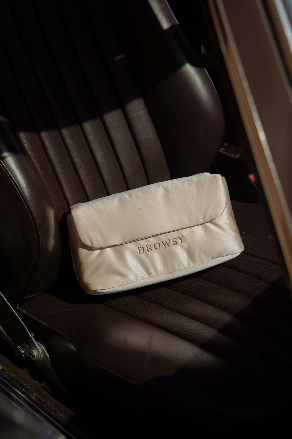 A cream-colored pouch with the word DROWSY embroidered in gold sits on the dark brown leather seat of a car.