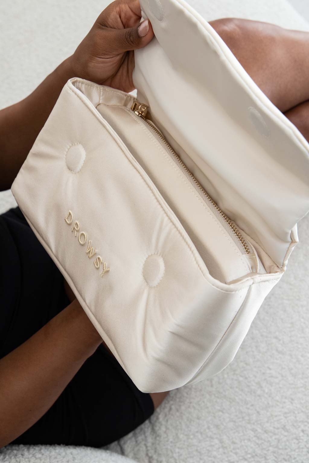 A person holds an open cream-colored bag with the word DROWSY embroidered on the front. The bag has a zipper and button details, and the background shows part of their crossed legs on a light surface.
