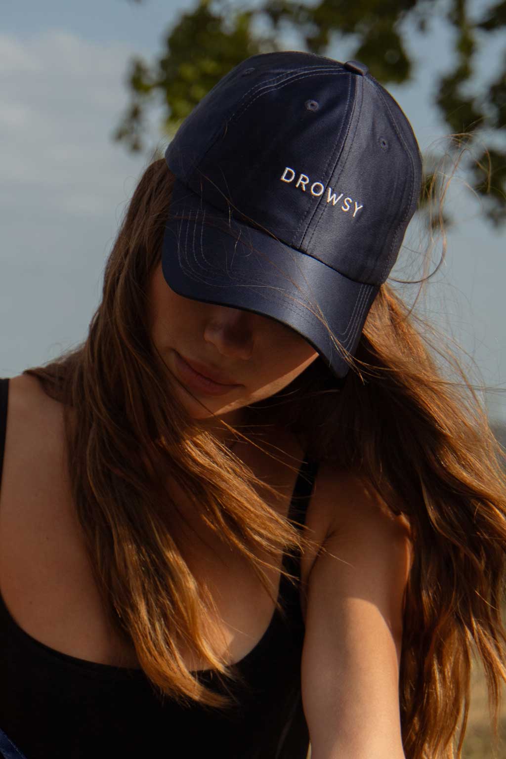 A person with long brown hair wears a navy blue cap with the word DROWSY embroidered in white letters, head slightly down, outdoors with trees and sky blurred in the background.